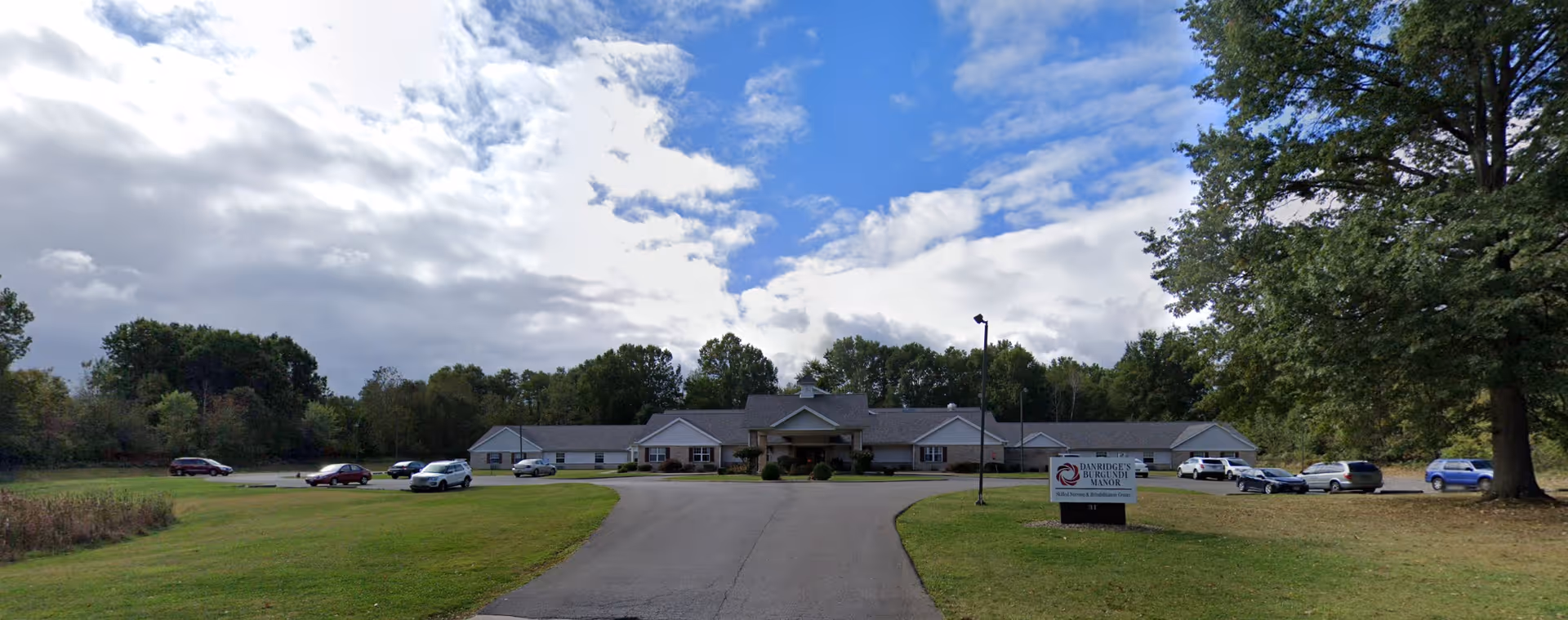 Wide exterior view of Danridge's Burgundi Manor, a single-story building with a gray roof surrounded by trees and a large grassy area. Several cars are parked in the parking lot in front of the building under a partly cloudy sky.