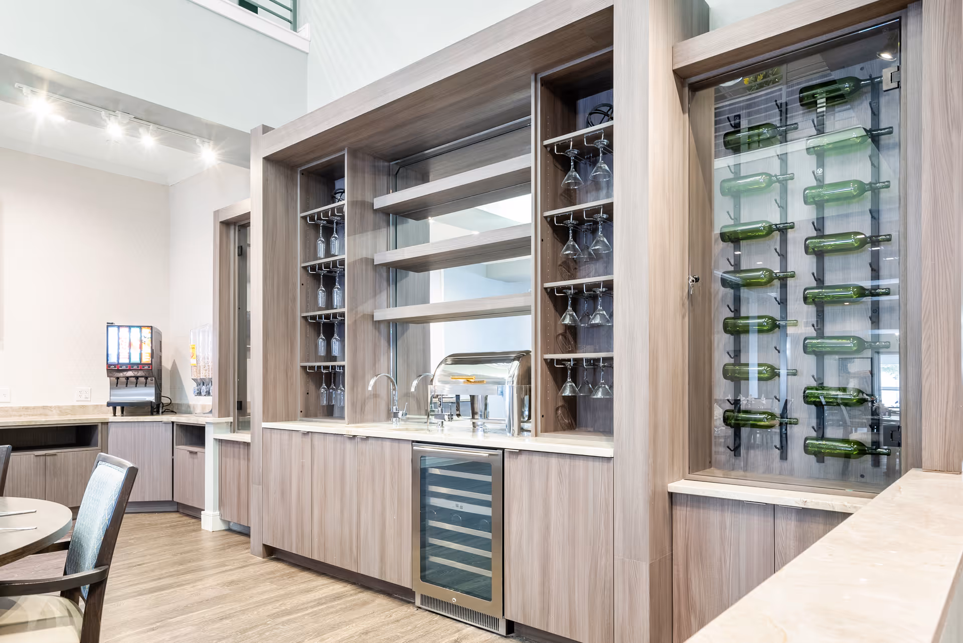 A modern dining area with a built-in wooden cabinet featuring glass shelves holding wine glasses and a wine bottle display. There is a small sink, a beverage cooler, and a drink dispenser in the background. The room has light-colored walls and wood flooring.