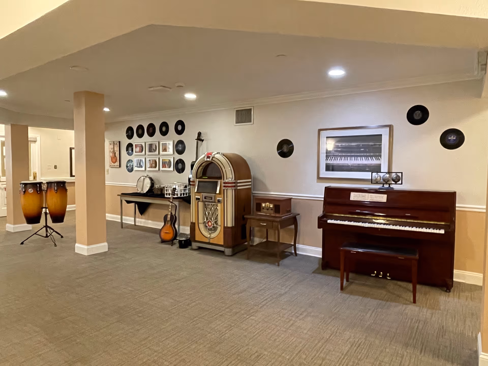 A music-themed common area in a senior living facility featuring a piano with a bench, a vintage jukebox, a small table with a retro radio, a guitar, a drum set, and framed pictures and vinyl records decorating the walls.