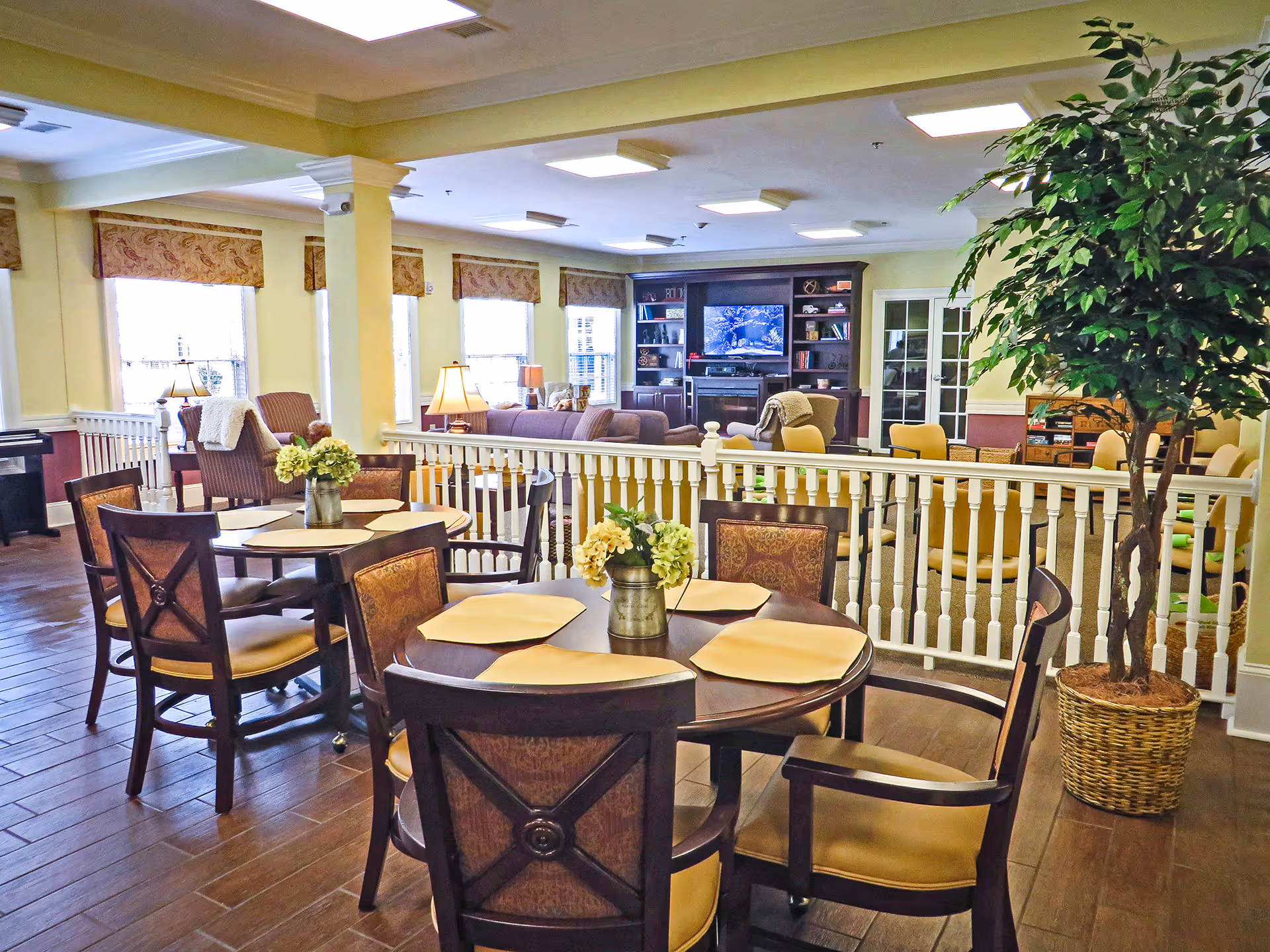 A bright and spacious common area in a senior living facility featuring round wooden tables with chairs, each table decorated with a small vase of flowers. The room has large windows with patterned valances allowing natural light to fill the space. In the background, there is a cozy seating area with armchairs and a large entertainment center with a TV. A large potted plant is placed near the railing that separates the dining area from the lounge.