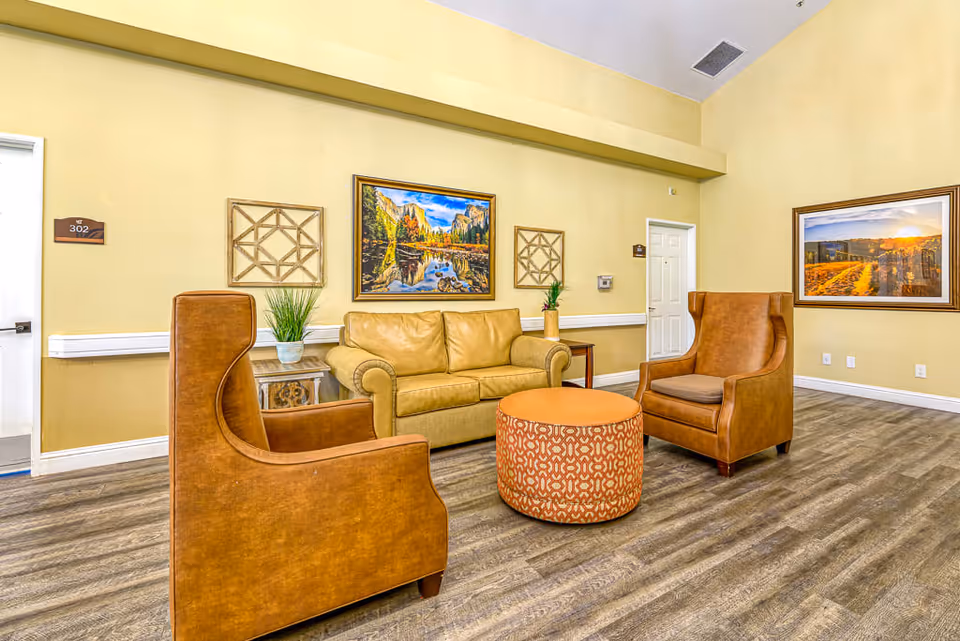 A cozy seating area in a senior living facility with two brown leather armchairs, a beige leather sofa, and a round orange patterned ottoman in the center. The walls are painted light yellow and decorated with framed landscape paintings and geometric wall art. The floor has wood-like vinyl planks, and there are two doors visible, one labeled 302.