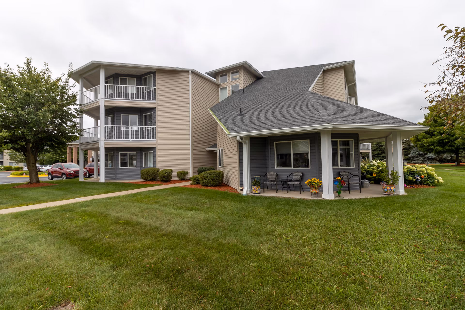 Exterior view of a multi-story senior living facility building with balconies, a covered patio area with chairs and potted plants, surrounded by green grass and trees under a cloudy sky.