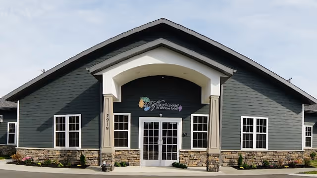 Front exterior view of Moraine Ridge Senior Living building with a peaked roof, multiple windows, stone facade on the lower half, and a double glass door entrance under a covered archway.