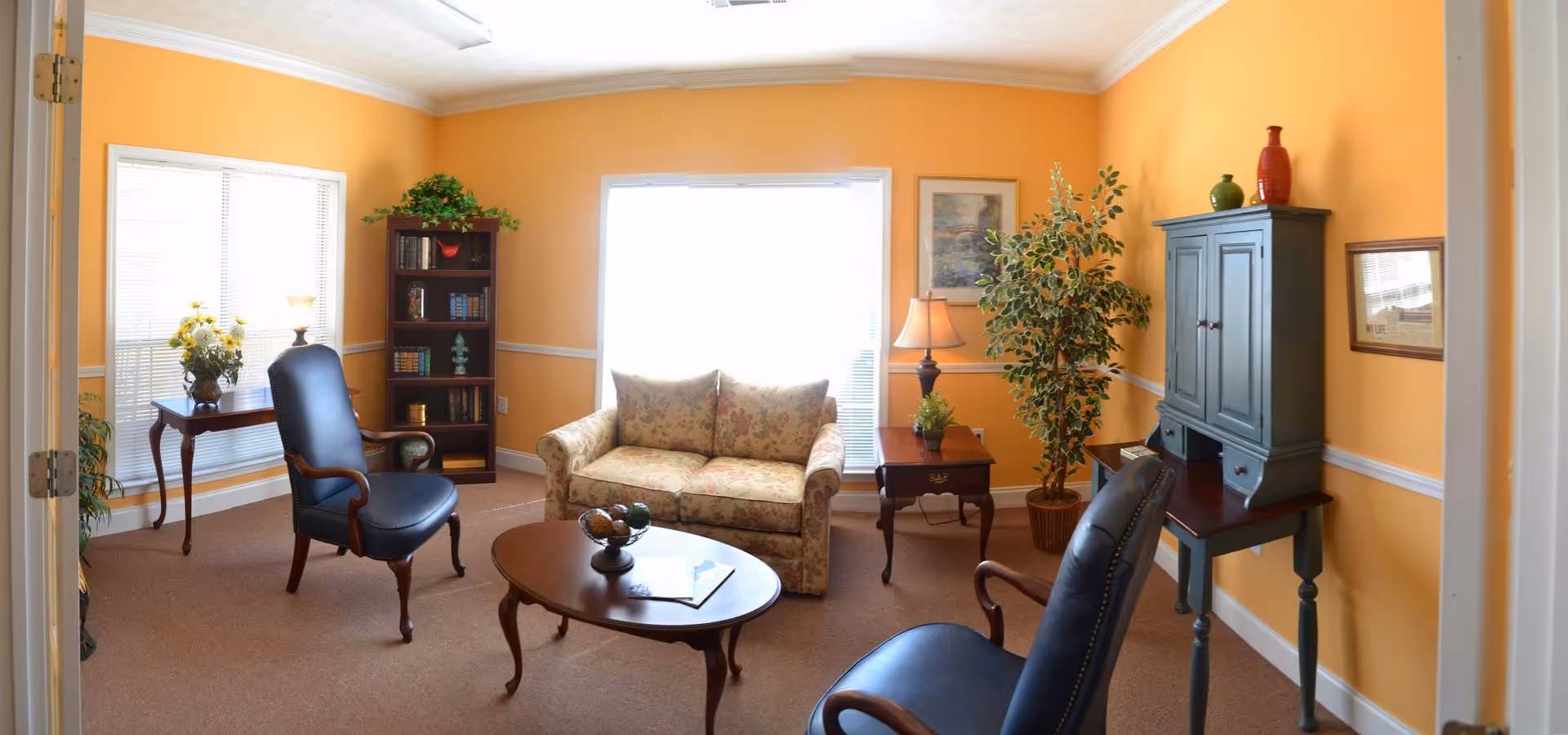 A cozy living room with warm yellow walls, featuring a floral-patterned loveseat, two dark leather armchairs, a wooden coffee table with decorative items, a wooden bookshelf filled with books, a side table with a lamp and plant, a small wooden desk with a cabinet, and large windows letting in natural light.
