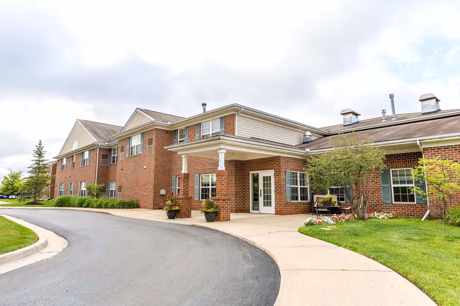 Exterior view of a two-story brick building with white trim and blue shutters, featuring a covered entrance with two large flower pots and a curved driveway leading up to it. There is a small garden with flowers and a bench near the entrance, and the sky is cloudy.