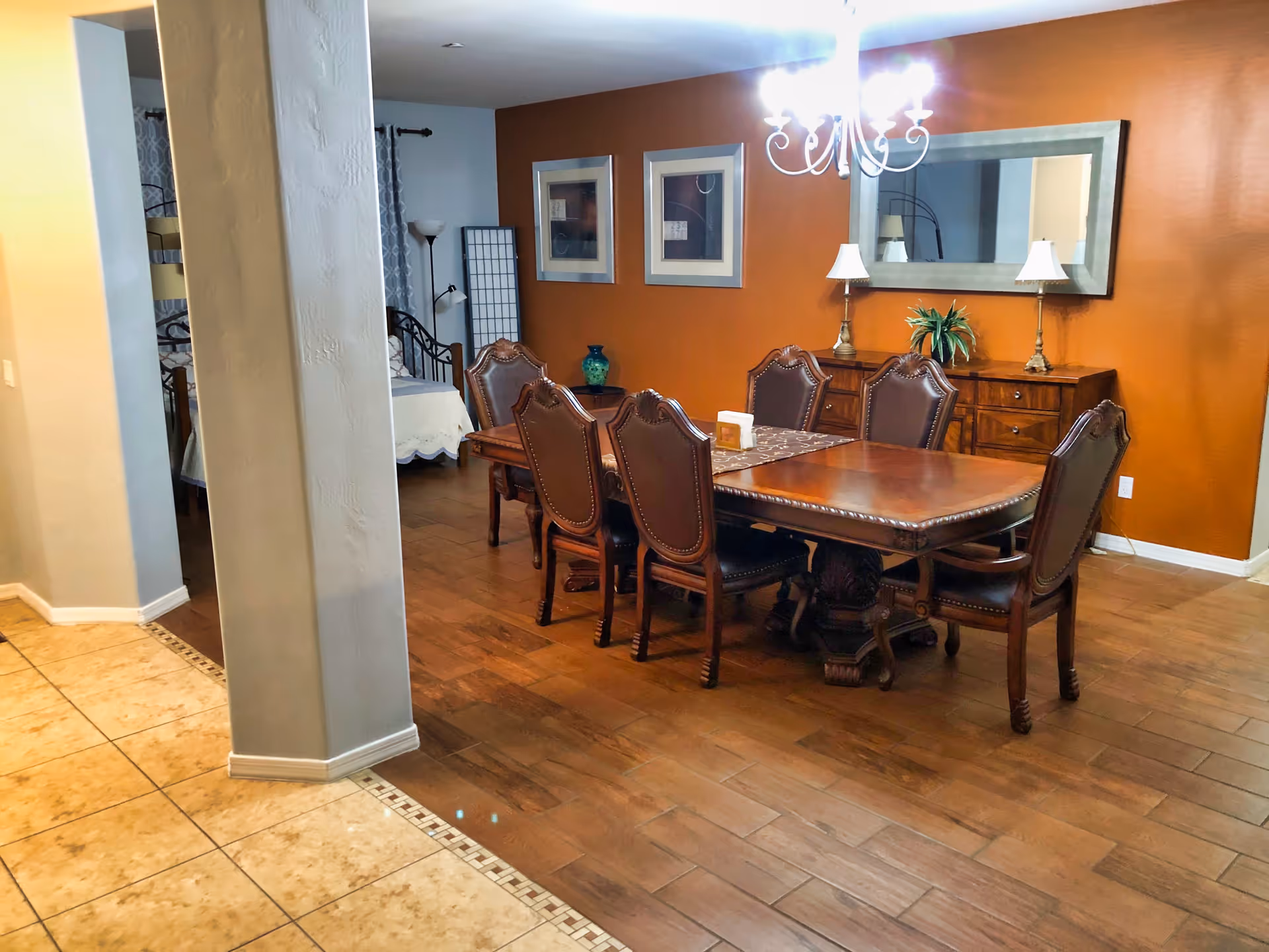 A dining room with a wooden dining table and six upholstered chairs. The room has a warm orange accent wall with a large mirror and two framed pictures. There is a chandelier hanging above the table, and a wooden sideboard with two lamps and a plant. The floor is wooden, and there is a tiled floor area adjacent to the dining space. In the background, a bedroom with a bed and a floor lamp is partially visible.