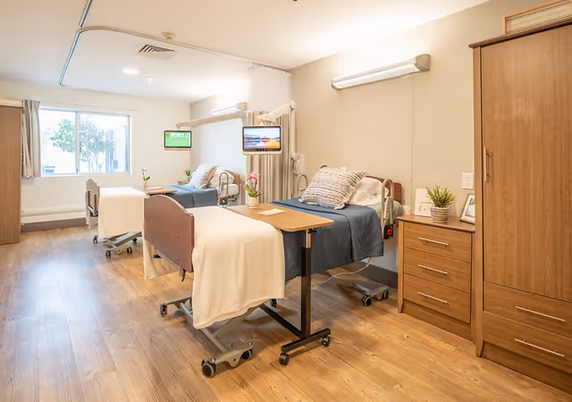 A bright and clean patient room with two hospital beds, each equipped with a blue blanket and white sheets. Each bed has an overbed table and a mounted television screen. The room features wooden flooring, a large window with curtains, a wooden dresser, and a wardrobe. There are small plants and framed pictures on the dresser, creating a welcoming atmosphere.