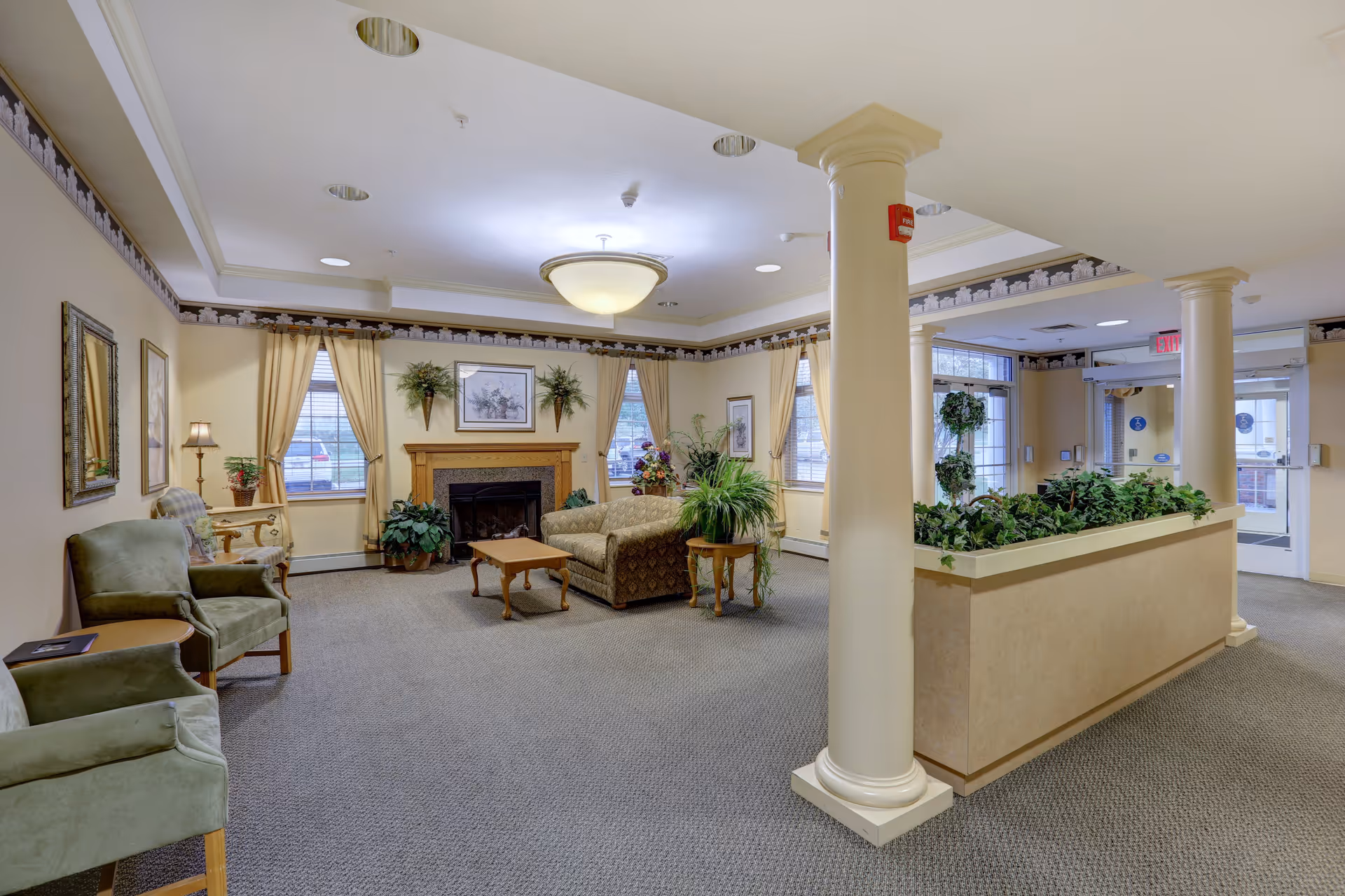 Well-lit senior living lounge with seating, a fireplace, indoor plants, and decorative columns near the entrance.
