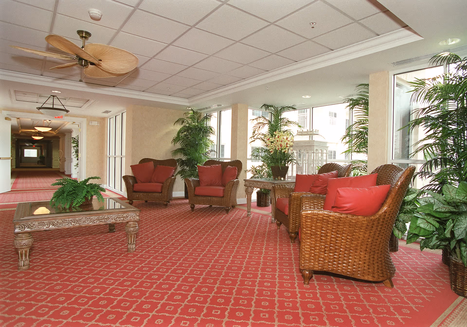 A bright and spacious indoor seating area with wicker chairs and a sofa featuring red cushions, a glass-top coffee table, and a side table with a flower arrangement. The area is decorated with several green plants and has large windows letting in natural light. The floor is covered with a red patterned carpet and the ceiling has white tiles with ceiling fans and light fixtures.