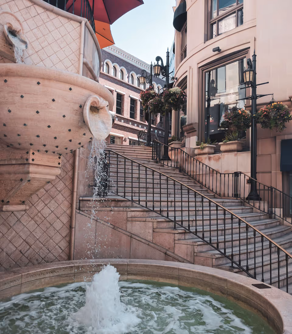 Stone fountain in front of a stepped plaza leading to an ornate building facade with hanging flower baskets and street lamps.
