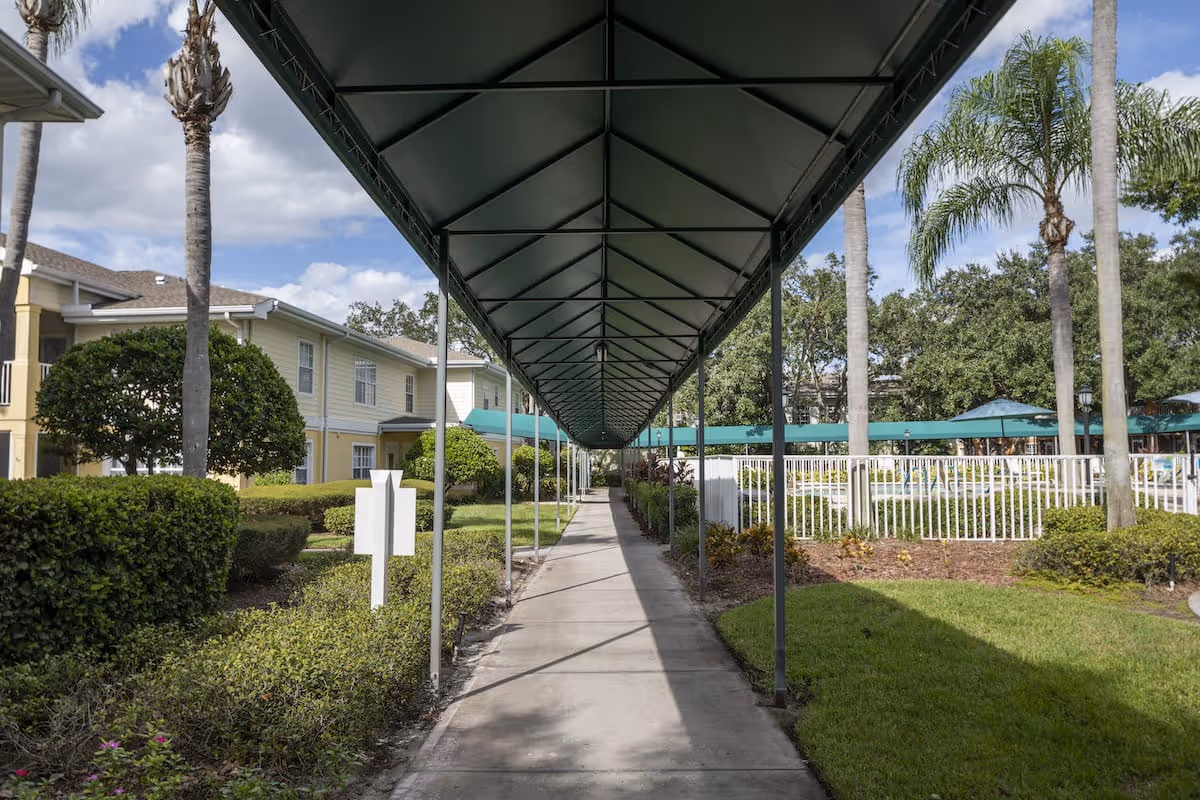 Covered outdoor walkway with a green canopy between landscaped gardens and residential buildings, with a fenced pool area visible to the right.