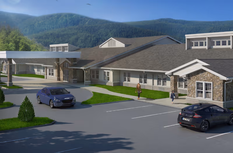Exterior view of Wynwood House senior living facility with a parking lot in front, two cars parked, and two people walking on the sidewalk. The building has a stone and siding facade with a covered entrance. Green hills and trees are visible in the background under a clear blue sky.