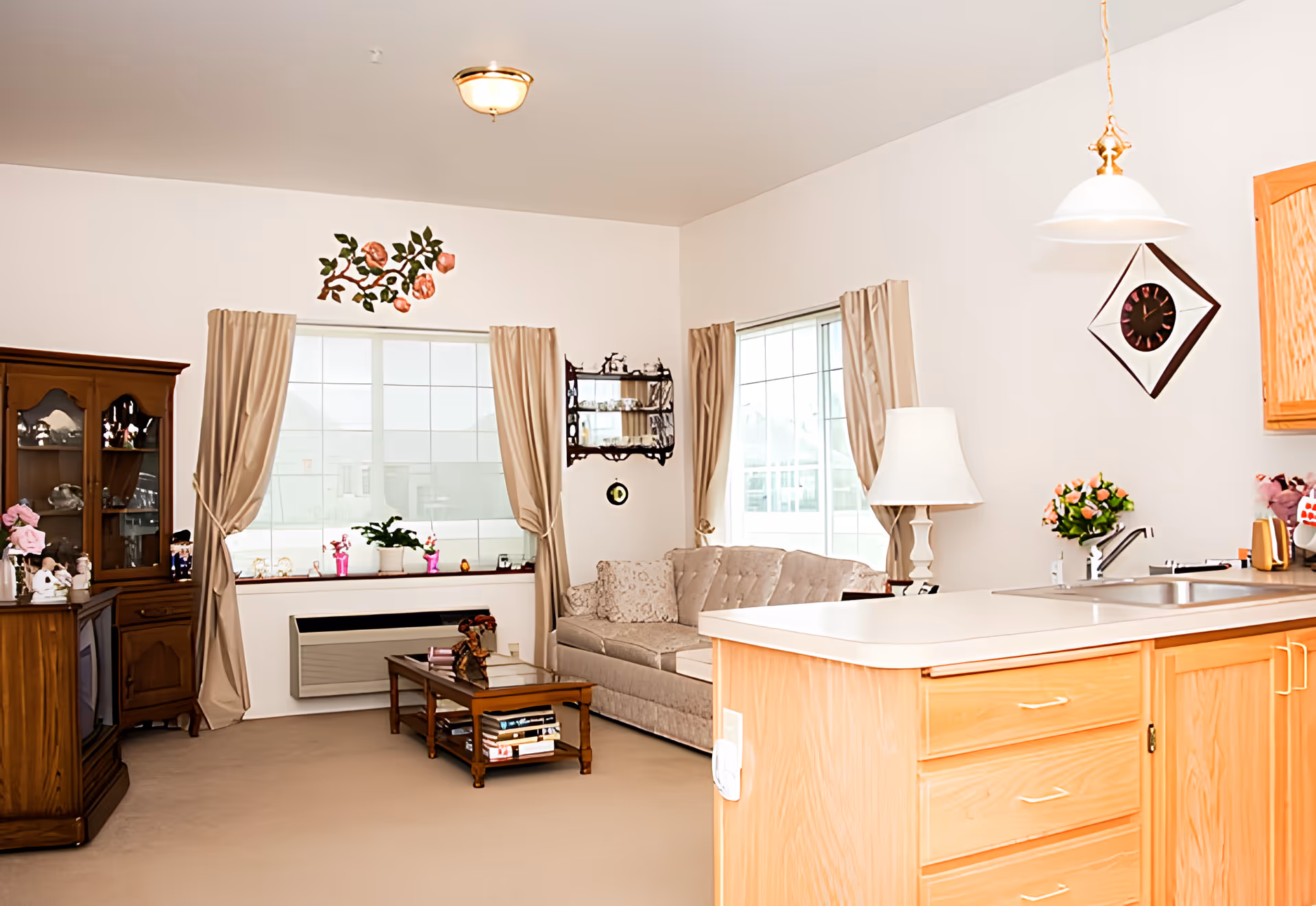 A cozy living room area with a beige couch, wooden coffee table with books, a wooden cabinet with glass doors, and a kitchen counter with a sink. The room has large windows with beige curtains, a wall clock, a table lamp, and decorative flowers on the counter and cabinet.