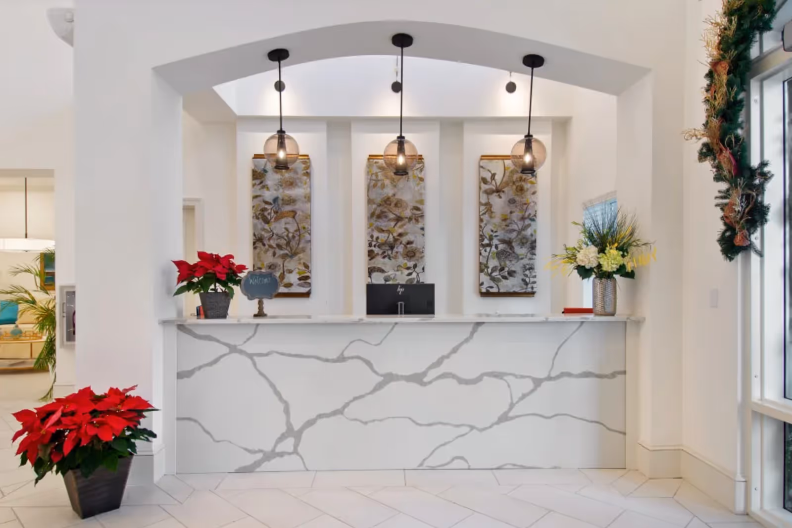 Reception area with a white marble-patterned front desk, three hanging pendant lights, floral artwork on the wall behind the desk, red poinsettia plants on the floor and desk, and a festive garland decoration on the right side near a window.