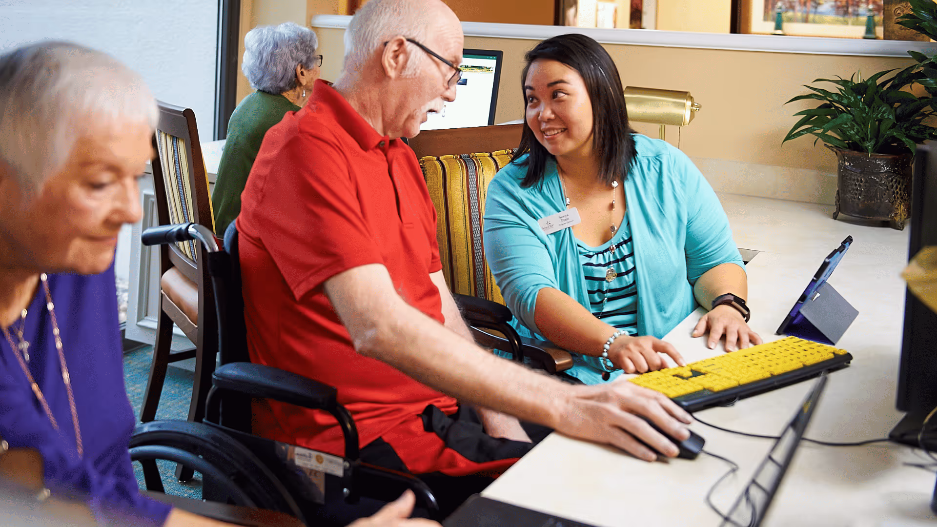 An elderly man in a wheelchair wearing a red shirt is using a computer with a large yellow keyboard, assisted by a smiling woman in a turquoise cardigan. Another elderly woman is seated nearby, and a fourth person is visible in the background.
