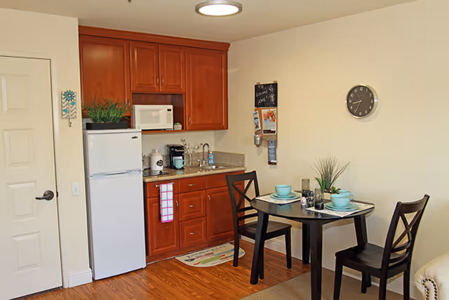 Small kitchenette with wooden cabinets, a refrigerator and microwave beside a round dining table set for two.