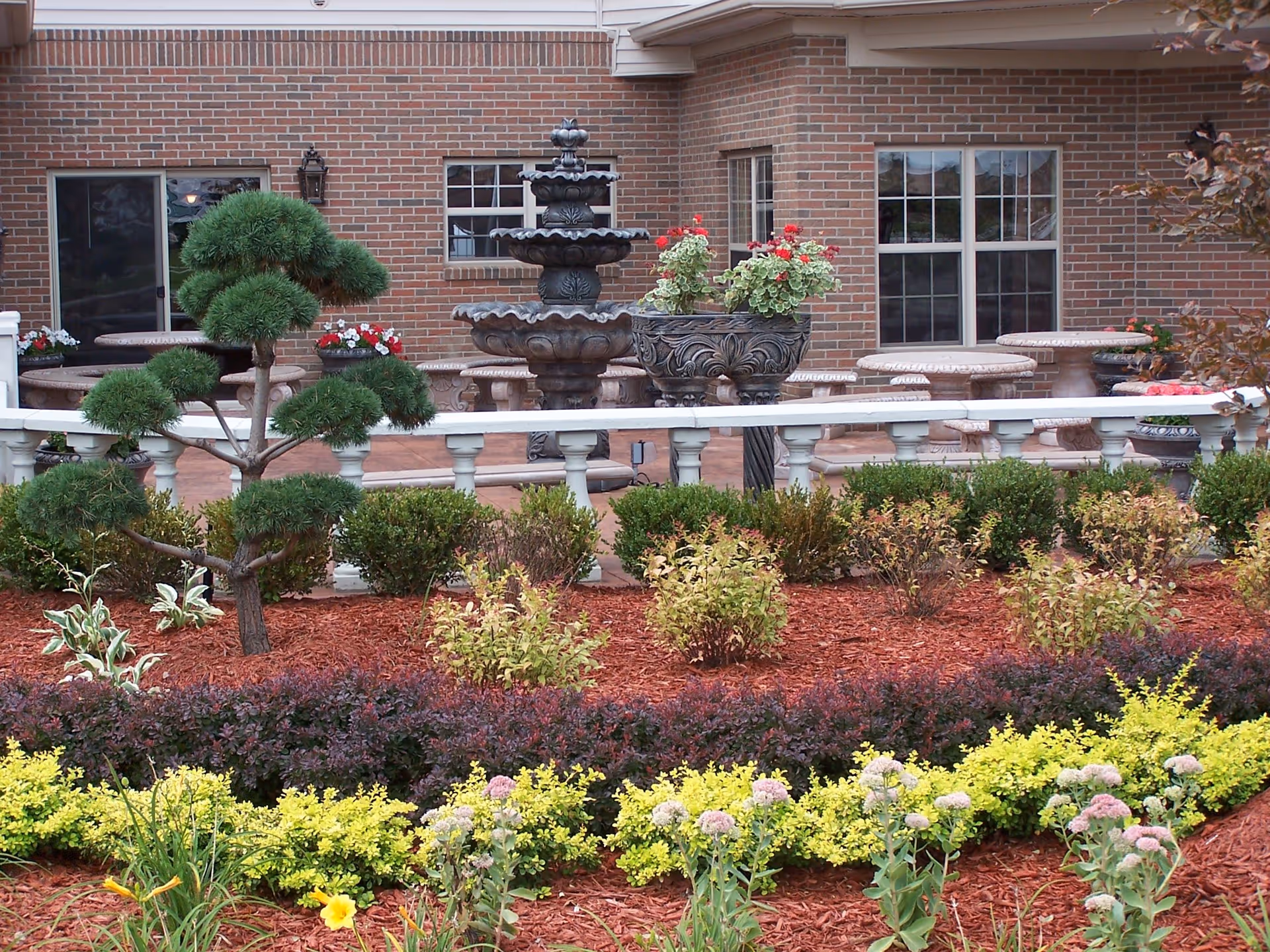 A landscaped garden area with various shrubs, flowering plants, and a small sculpted tree in front of a brick building. There is a white railing separating the garden from a patio area with stone tables and benches, and a multi-tiered water fountain is visible on the patio.
