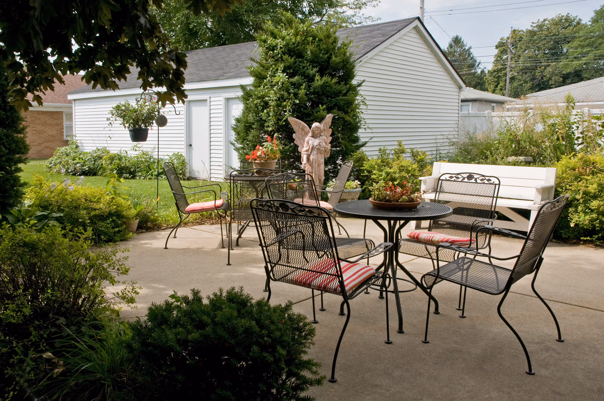 An outdoor patio with wrought-iron table and chairs, potted plants, a decorative angel statue, and a white shed in the background.