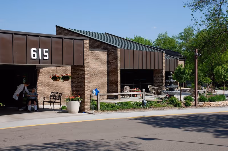 Exterior view of a brick building with the number 615 displayed above the entrance. There is a covered walkway with a person in a white coat attending to an elderly person in a wheelchair near a bench. The area has potted plants, hanging flower baskets, outdoor seating, and a multi-tiered fountain surrounded by greenery under a clear blue sky.