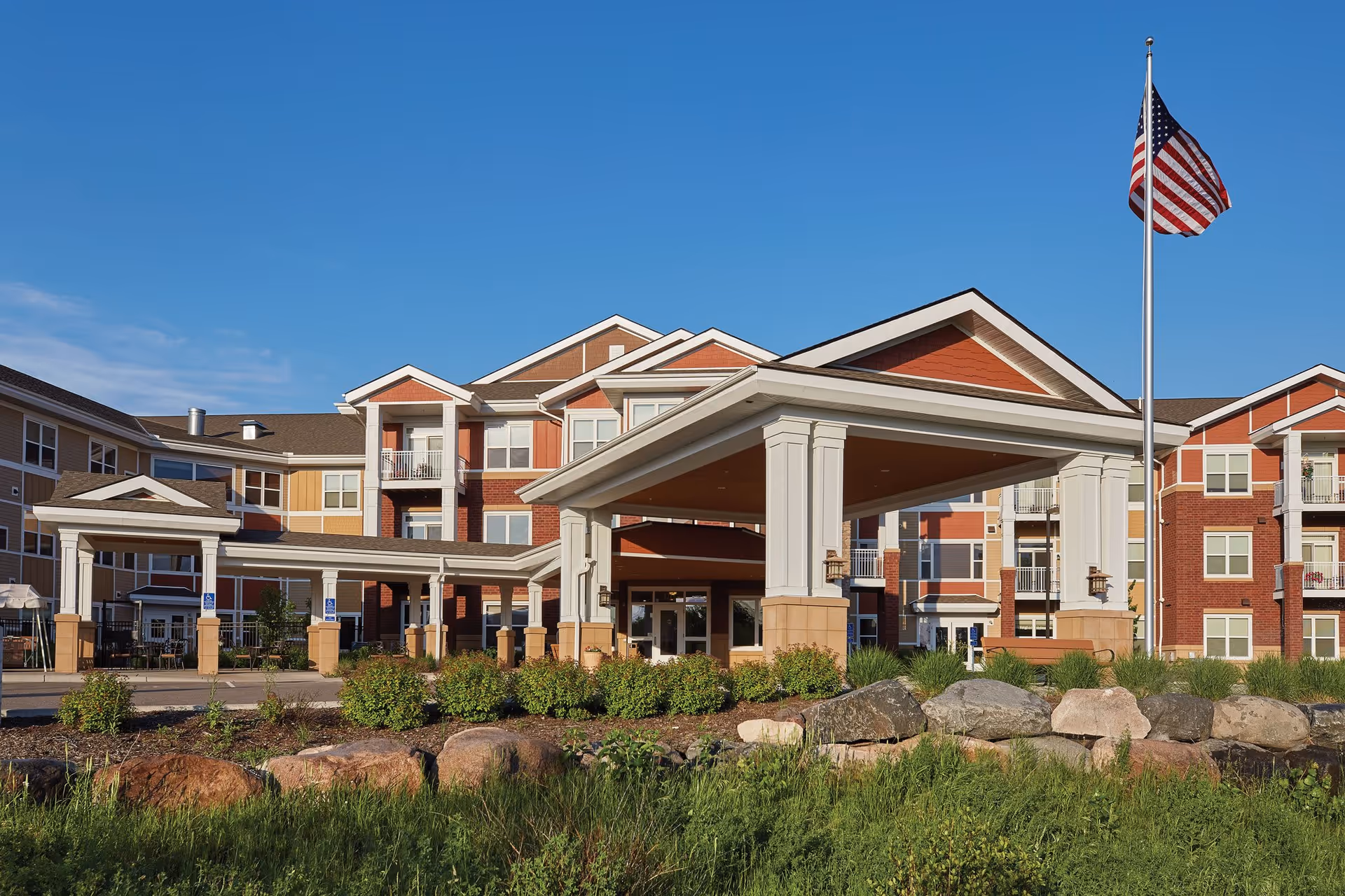 Exterior view of a multi-story senior living facility with a covered entrance, landscaped greenery, and an American flag on a flagpole against a clear blue sky.