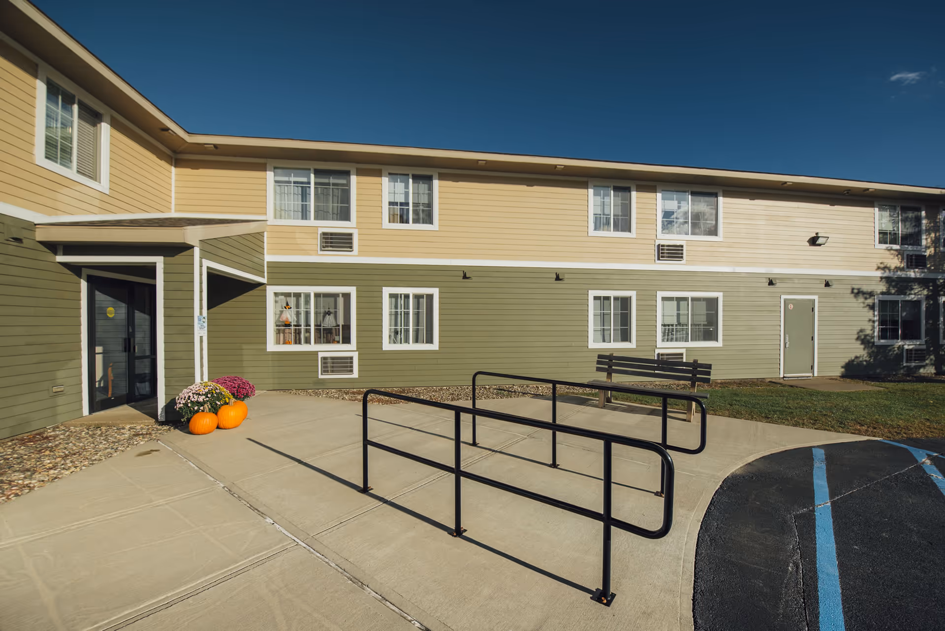 Exterior view of a two-story senior apartment building with a front entrance, accessible ramp, and pumpkins by the doorway.