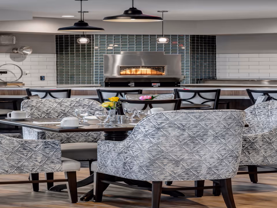 A modern dining area with patterned upholstered chairs around a wooden table set with glassware, plates, and a small vase with yellow and pink flowers. In the background, there is a pizza oven built into a tiled wall with pendant lights hanging from the ceiling.