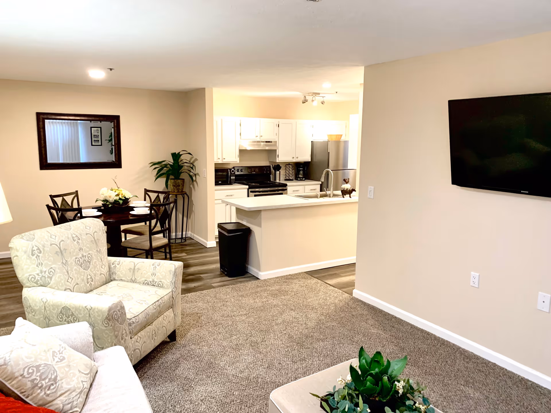 Interior view of a senior living facility showing a living room with a patterned armchair and sofa, a wall-mounted flat-screen TV, a small dining area with a round table and four chairs, and a kitchen with white cabinets, a stove, refrigerator, and a countertop with a sink. There is a plant near the dining area and a mirror on the wall.