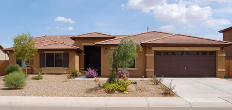Single-story house with a brown tiled roof, beige exterior walls, and a two-car garage. The front yard features desert landscaping with gravel, small bushes, and a few trees under a partly cloudy blue sky.
