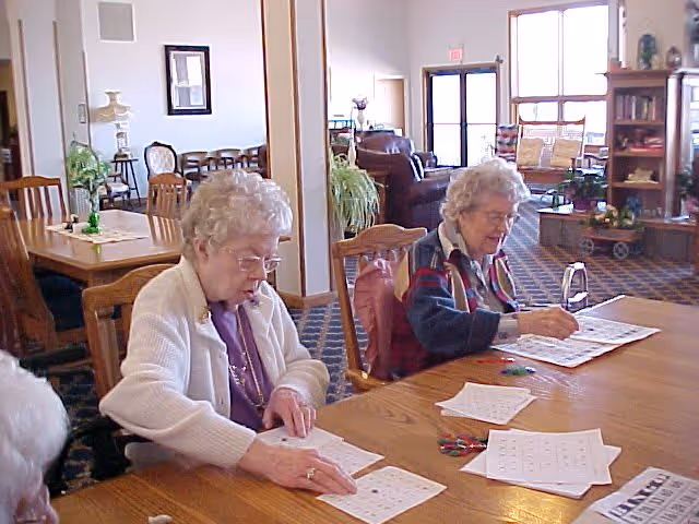 Two elderly women sitting at a wooden table in a common area, engaged in playing bingo with bingo cards and markers spread out on the table. The room has carpeted floors, wooden furniture, plants, and large windows letting in natural light.
