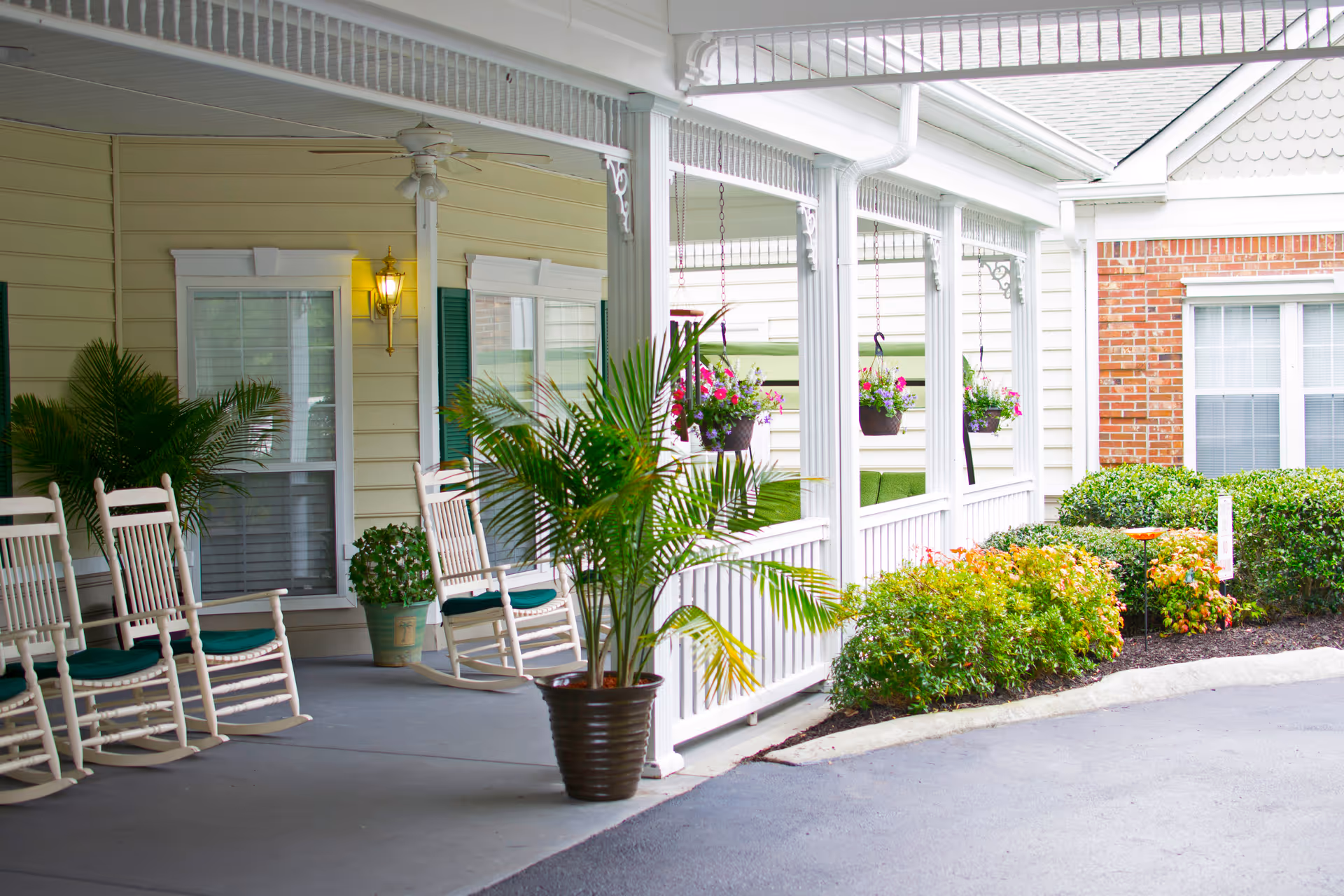 Covered porch area with white rocking chairs, potted plants, hanging flower baskets, and a yellow exterior wall with windows and a lit wall-mounted lantern.