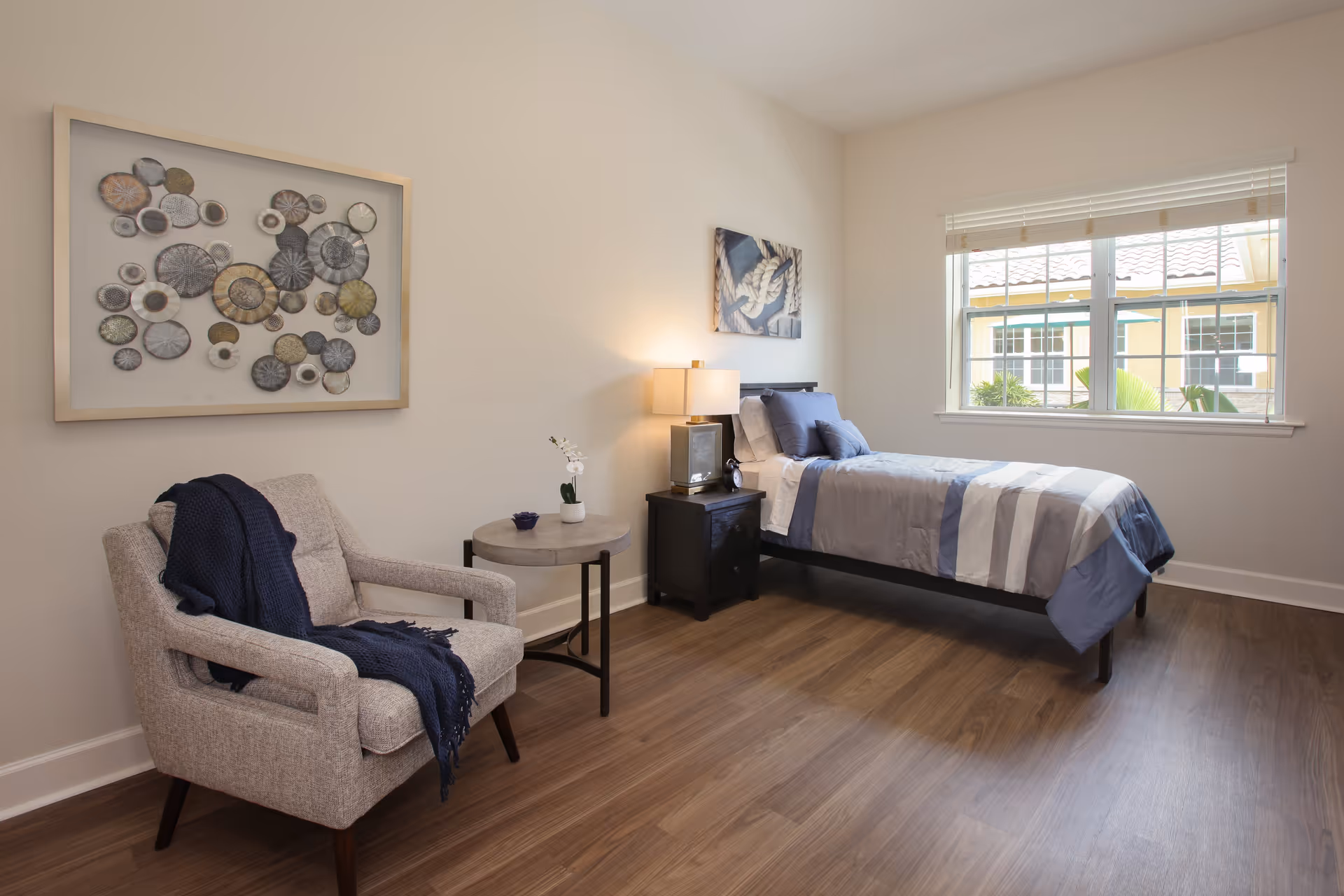 Bright, clean bedroom with a single bed beside a nightstand and lamp, an upholstered armchair with a throw, decorative wall art, and a window.