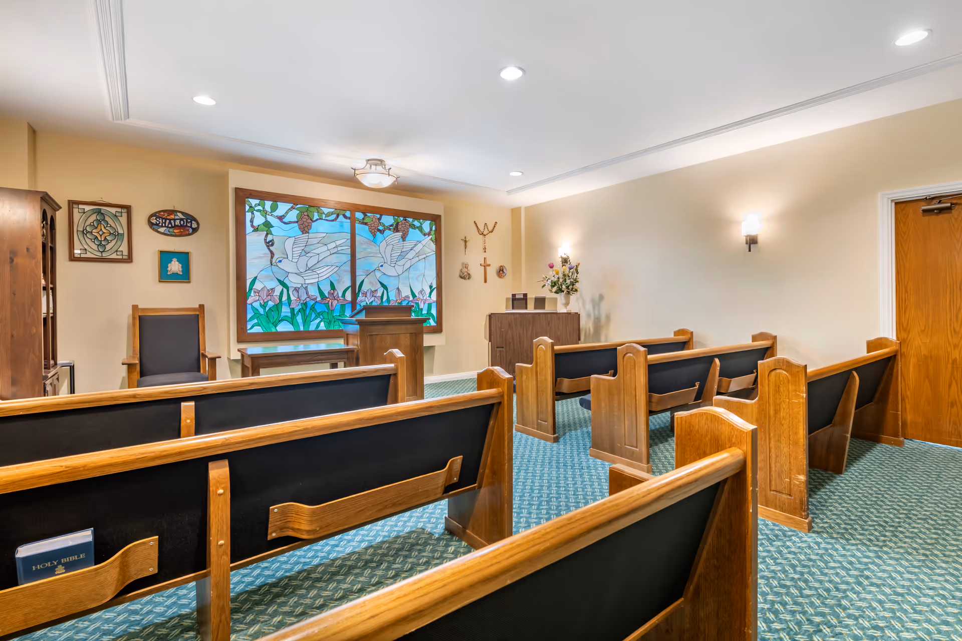 Small chapel interior with wooden pews, a pulpit, and a large stained-glass window depicting doves.