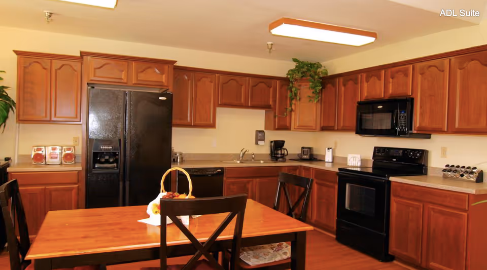 A kitchen area with wooden cabinets, a black refrigerator, black stove, black microwave, and a wooden dining table with chairs. There are small appliances like a coffee maker and toaster on the countertop, and a basket with fruit on the table. A plant is placed on top of the cabinets.