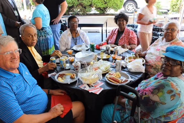 A group of elderly people sitting around a round table outdoors, enjoying a meal together. The table is covered with a black tablecloth and has plates of food, drinks, and bowls with salad and other dishes. Some people are smiling and engaging with each other, and there are trees and a parked car visible in the background.