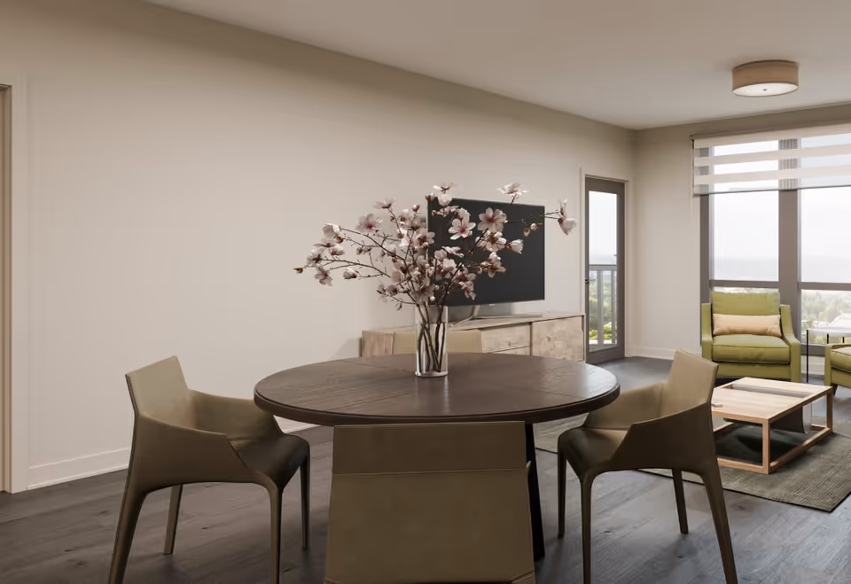 A modern living and dining area with a round wooden dining table and four chairs. A vase with pink flowers is placed on the table. In the background, there is a TV on a wooden cabinet, two green armchairs with a small wooden coffee table between them, large windows with blinds, and a door leading to a balcony with a scenic view.