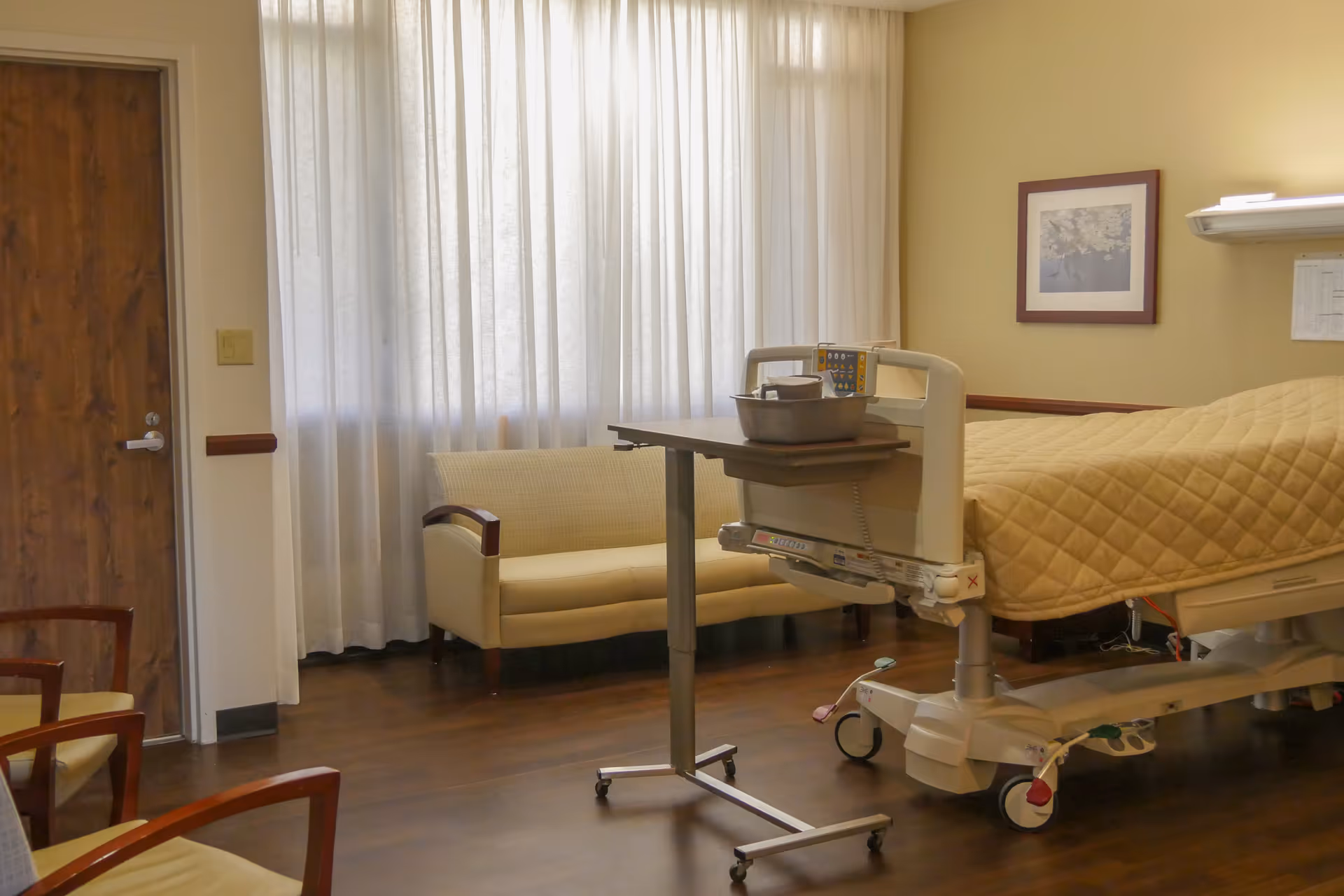 A hospice patient room with a hospital bed covered in a beige quilt, an overbed table with medical supplies, a beige loveseat near a window with sheer curtains, and two wooden chairs with beige cushions. The room has wooden flooring, a wooden door, and a framed picture on the wall.
