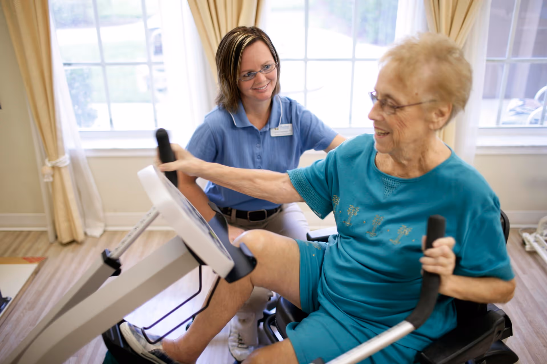 An elderly woman in a teal outfit exercises on a seated elliptical machine while a caregiver in a blue shirt and glasses smiles and supports her in a bright room with large windows and beige curtains.