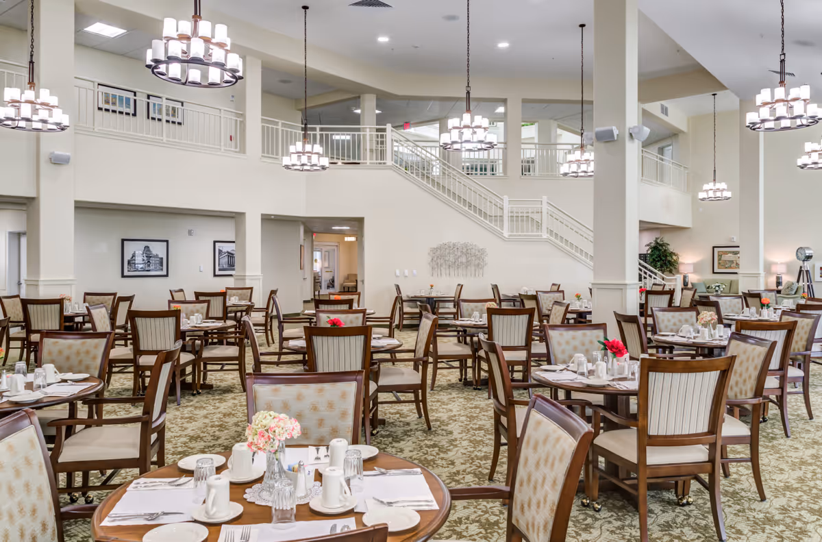 Spacious dining room in Bella Vista Gracious Retirement Living with multiple round wooden tables set with white plates, cups, and silverware. The room features high ceilings with hanging chandeliers, beige walls, carpeted floor, and a staircase leading to an upper level. The decor includes framed pictures on the walls and small flower arrangements on the tables.