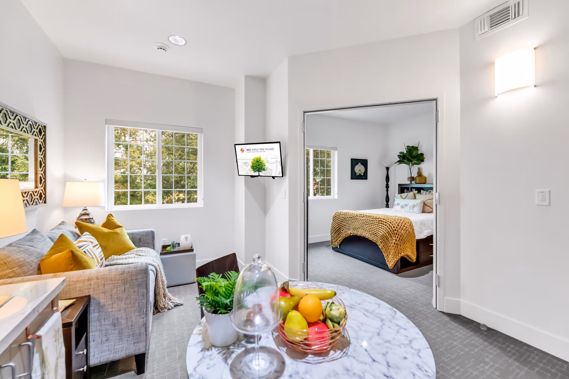 A bright and modern assisted living apartment featuring a small living area with a gray couch adorned with yellow and patterned pillows, a marble-top round table with a fruit basket and a small plant, and a bedroom visible through double doors with a bed covered in white linens and a mustard-colored knitted throw. The room has large windows letting in natural light, a wall-mounted TV displaying ACC Maple Tree Village branding, and neutral-colored walls and carpet.