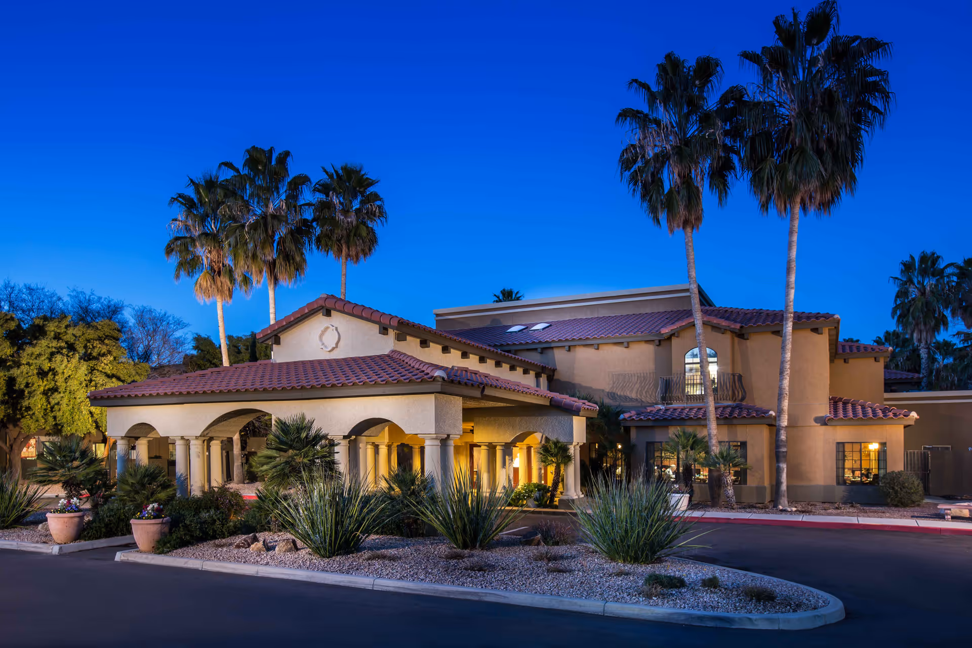 Exterior view of a senior living facility building at dusk with warm lighting, palm trees, desert landscaping, and a clear blue sky.
