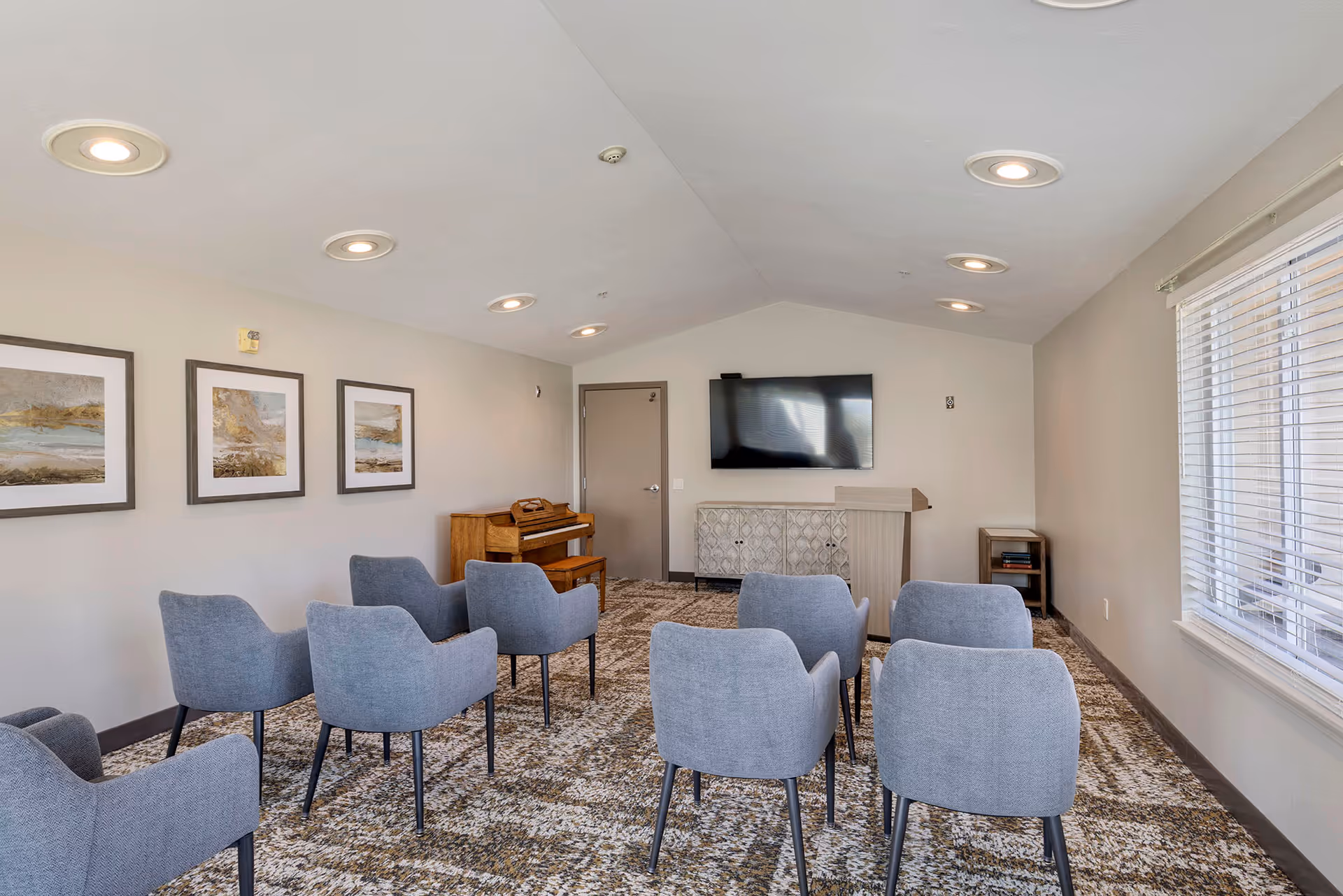 Small activity room with rows of gray chairs facing a wall-mounted TV, a piano, and framed artwork on the wall.