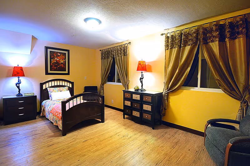 A cozy bedroom with a single dark wooden bed featuring a colorful bedspread and pillow. There are two windows with gold curtains, a dark wooden dresser with a red lamp on top, a matching nightstand with another red lamp, a framed floral painting on the wall, and a gray armchair in the corner. The room has wooden flooring and warm yellow walls.