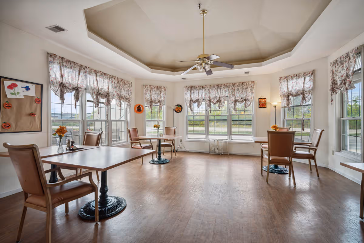 A bright dining room with multiple tables and chairs arranged around the room. The room features large windows with floral curtains, a ceiling fan with lights, and wooden flooring. Small flower vases are placed on the tables, and there is a bulletin board with decorations on one wall.