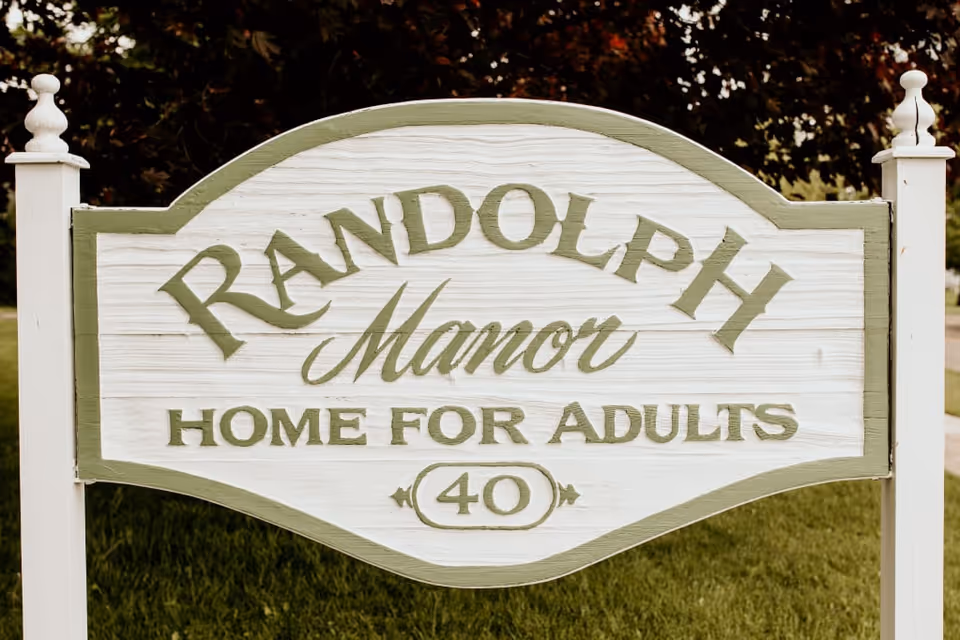 Wooden sign on posts reading "Randolph Manor Home for Adults 40" set on grass with trees in the background.
