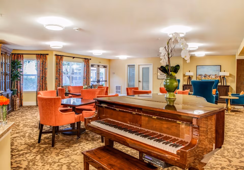 Bright senior living common room with a polished grand piano in the foreground, orange upholstered chairs and tables, and large windows.