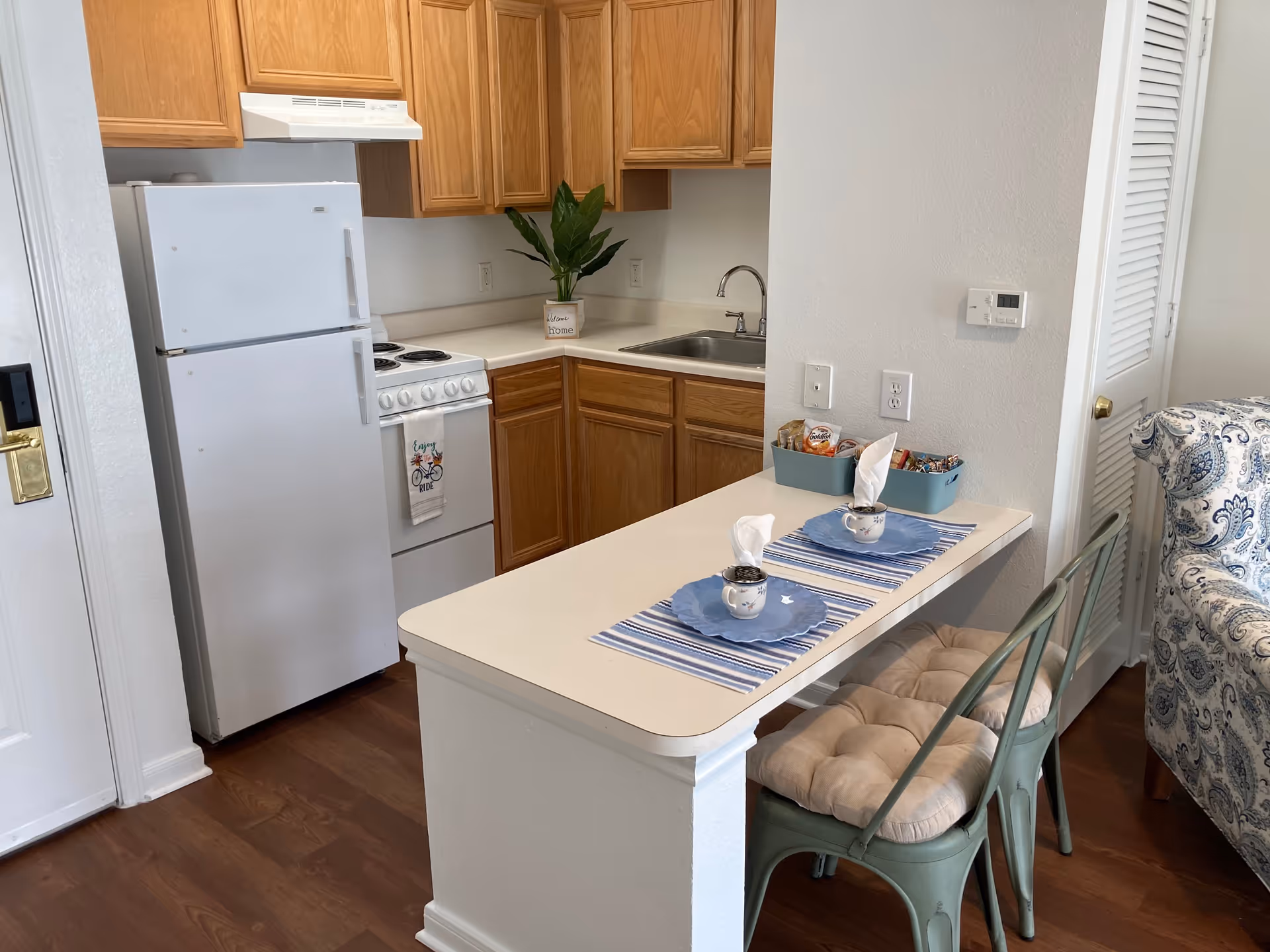 Small kitchen area with wooden cabinets, white refrigerator, stove, and sink. A narrow counter extends outward with two place settings including blue plates, cups with napkins, and striped placemats. Two cushioned metal chairs are placed at the counter. Part of a patterned upholstered chair is visible on the right side.