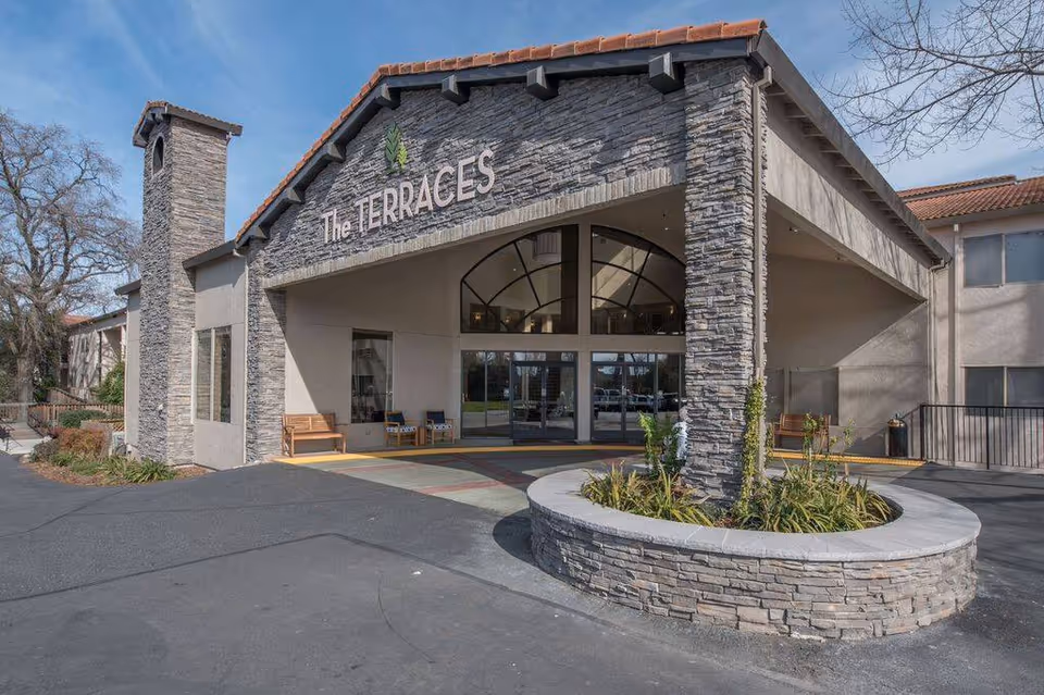 Covered stone-clad entrance of a senior living building with a circular planter and 'The TERRACES' sign above the driveway.