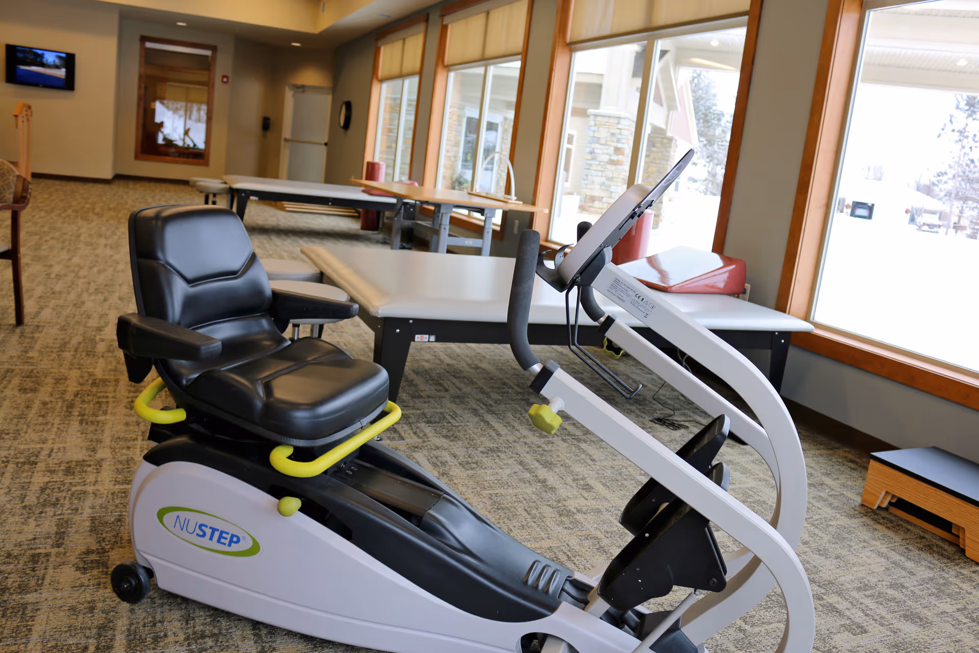 An indoor therapy room with a NuStep recumbent exercise machine positioned in front of large windows.