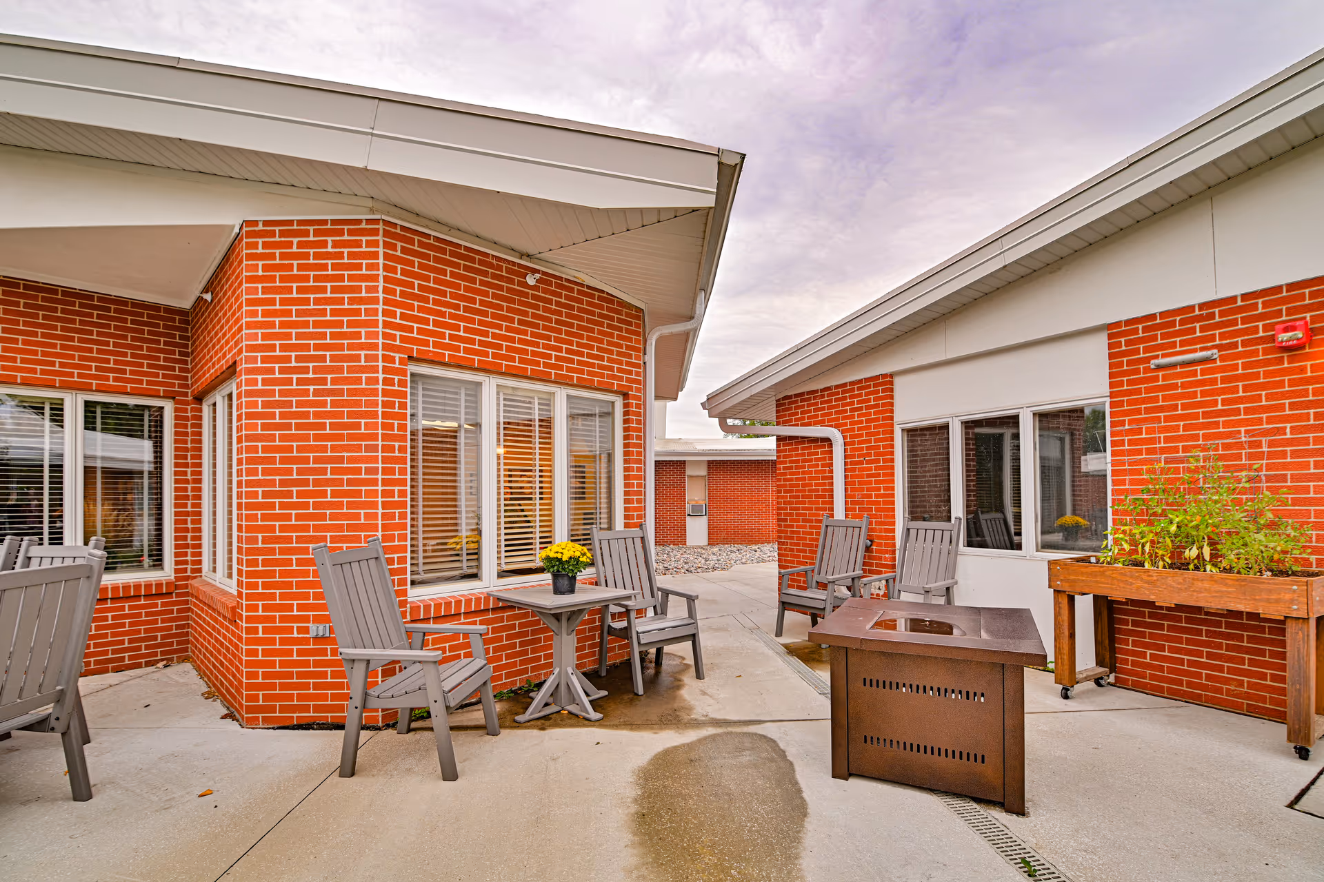Outdoor patio area at Valley Senior Living on Cherry - Tufte Manor featuring red brick buildings, gray patio chairs, a small table with a potted plant, a fire pit, and a raised garden bed under a cloudy sky.