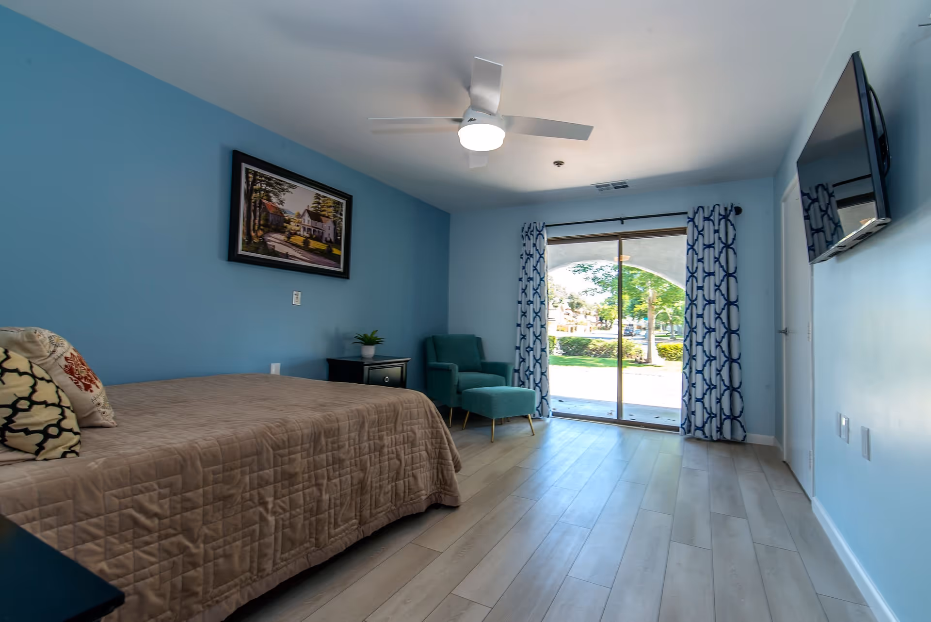 A bright bedroom with light blue walls and light wood flooring. The room features a bed with a beige quilt and decorative pillows, a black nightstand with a small plant, a teal armchair with an ottoman, a ceiling fan with a light, a wall-mounted flat-screen TV, and large sliding glass doors with patterned blue and white curtains that open to a green outdoor area.